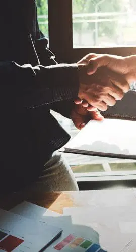 Two individuals are shaking hands over a table with documents and a digital tablet, suggesting a professional agreement or partnership.