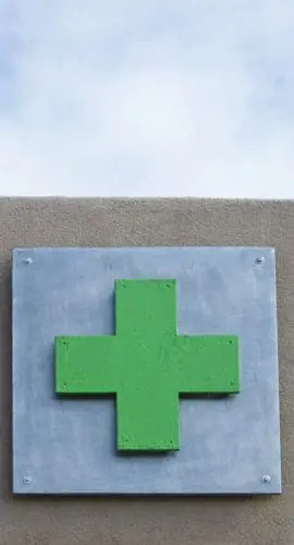 A green cross sign, which often represents a pharmacy or medical services, is mounted on a wall under a blue sky with clouds.