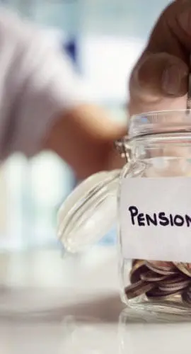 A hand is placing coins into a glass jar labeled "PENSION" on a table.