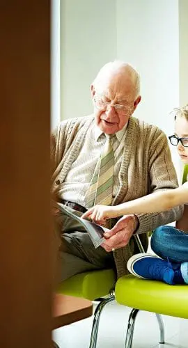 An elderly man and a young boy are sitting on a green bench, with the man showing something to the boy from a book or a tablet.