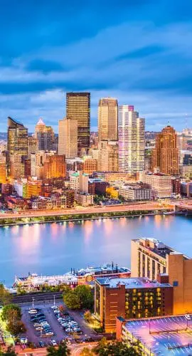 A panoramic twilight view of a city skyline with illuminated buildings reflected on a calm river.