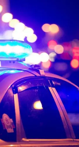 A police car with flashing blue lights is parked at night with blurred city lights in the background.