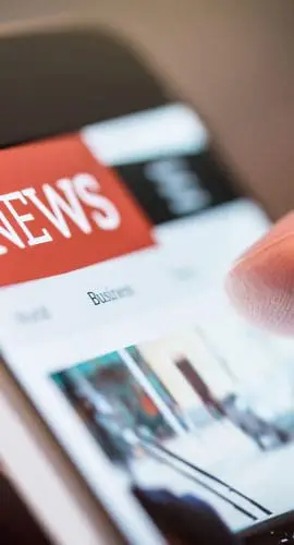A close-up view of a person's hand holding a smartphone displaying a news website with a red "NEWS" banner at the top.