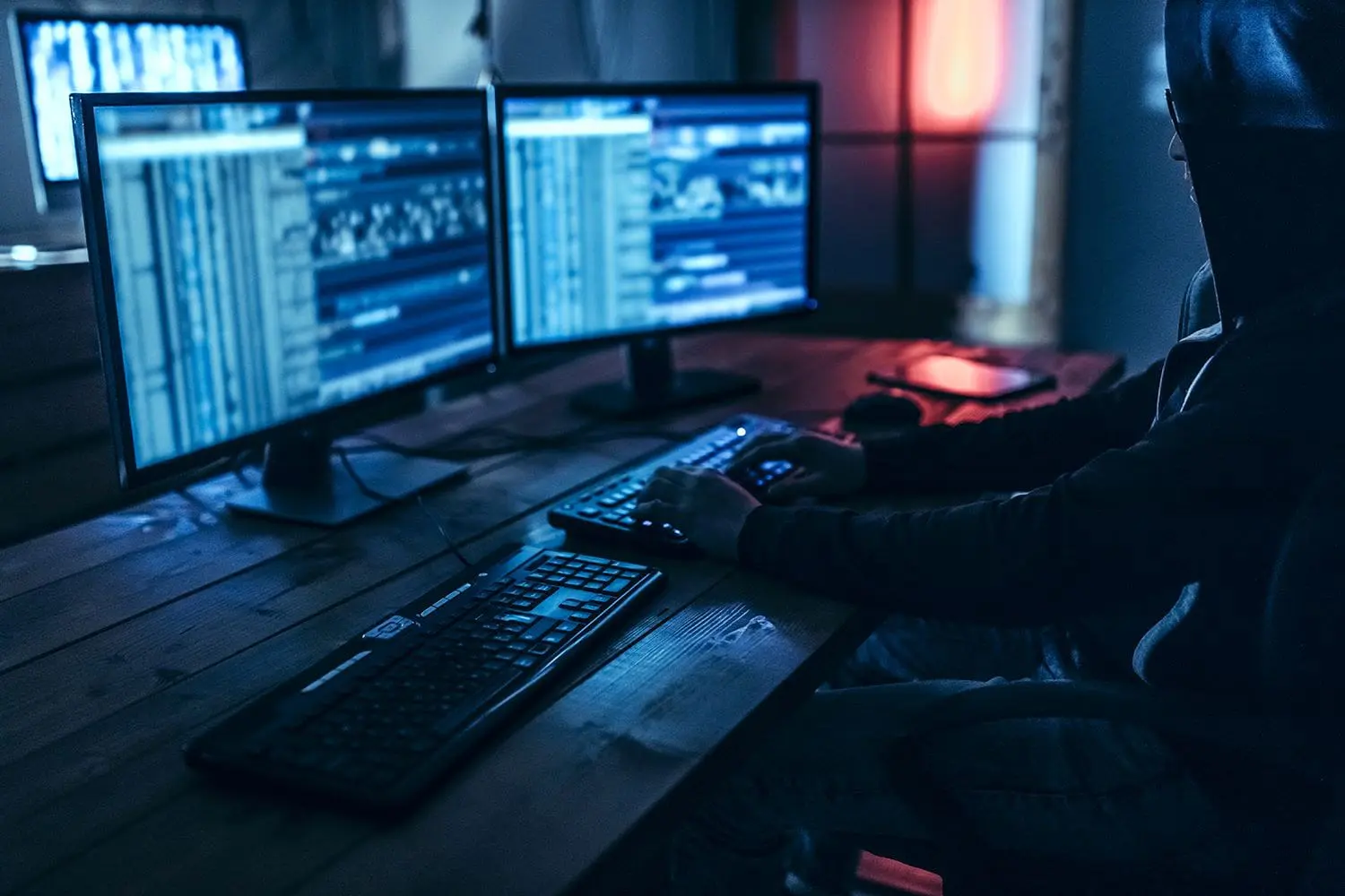 A silhouette of a person is working at a desk with multiple computer monitors displaying code or data in a dimly lit room with a blue hue.