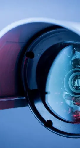 A close-up of a security camera with a red recording light, mounted on a wall against a blue background.