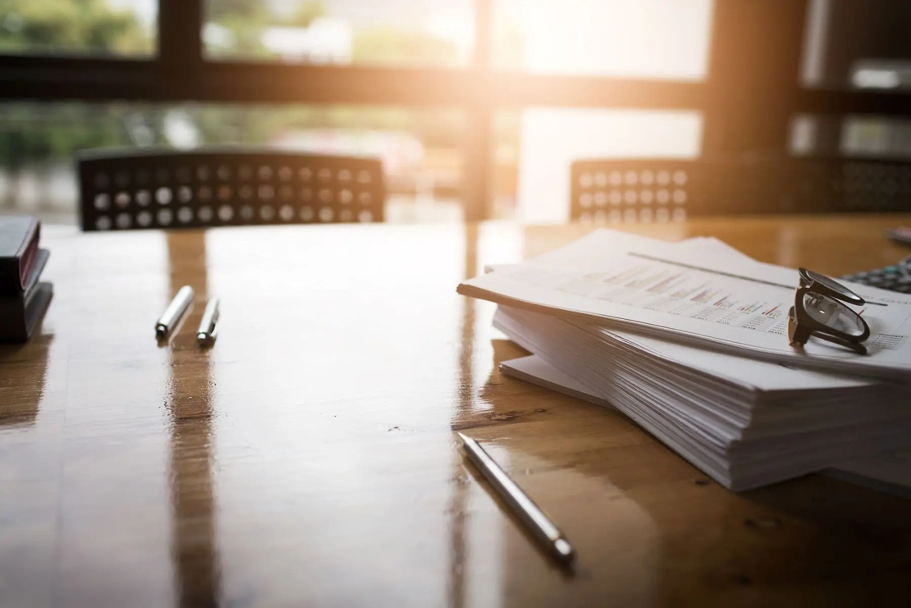 A stack of documents with a pen on top rests on a wooden table in a warmly lit room, suggesting a workplace setting with a focus on the paperwork.