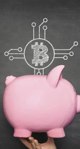 A hand holds a pink piggy bank in front of a blackboard with a Bitcoin symbol and circuit lines drawn on it.