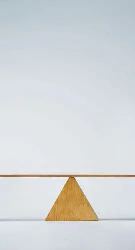 A piggy bank and a jar of pills are balanced on a wooden seesaw against a plain background.
