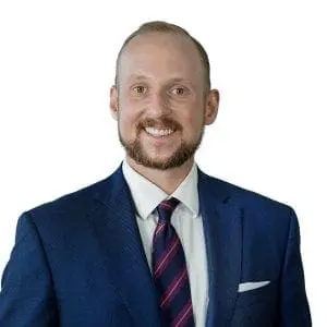 A man in a blue suit, striped tie, and white shirt is smiling against a white background.
