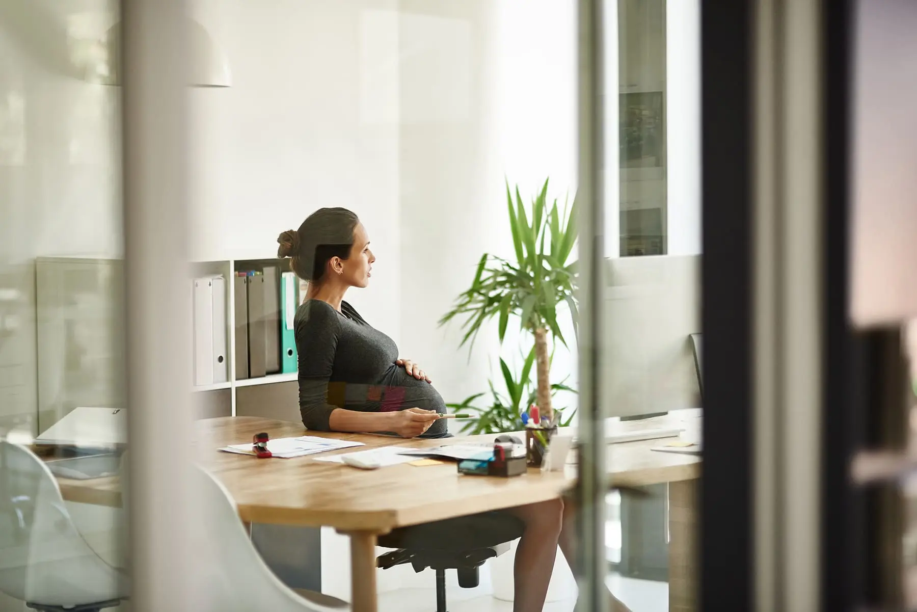 A pregnant woman is sitting at her office desk, holding her belly while gazing out the window.