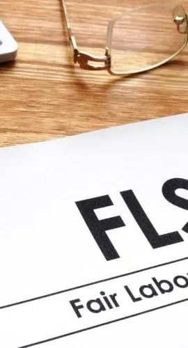A document titled "FLSA: Fair Labor Standards Act" is placed on a wooden desk next to a pair of glasses and calculator keys.