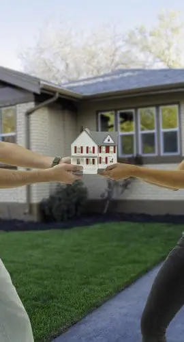 Two people are pulling on a small model house while standing in front of a suburban home.