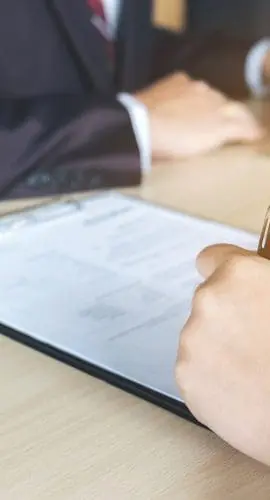 Two individuals are sitting at a table, one using a laptop and the other writing on a clipboard.