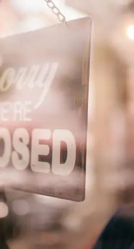 A person is standing inside, looking out a window with a "Sorry, we're closed" sign hanging in front.