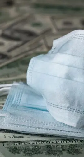 Two surgical masks are placed on top of numerous U.S. dollar bills.