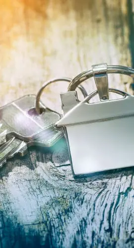 House keys with a keychain shaped like a house lie on a rustic wooden surface.