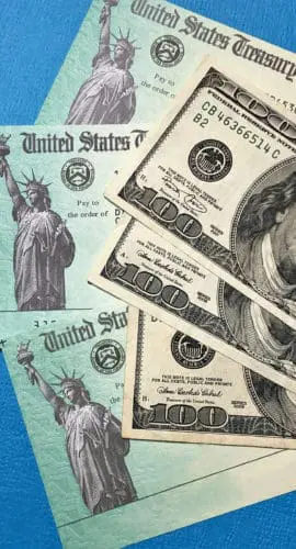 Several 100-dollar bills are placed on top of documents that appear to be U.S. Treasury checks.