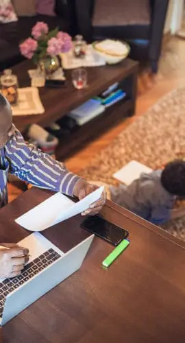 A person works on a laptop at a table while two children draw on the floor nearby.