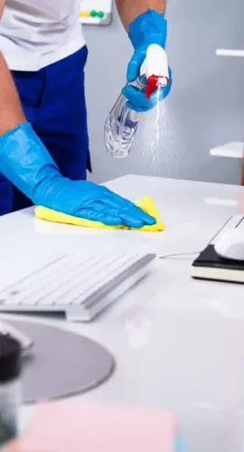 A person wearing blue gloves is cleaning a desk with a spray bottle and cloth.