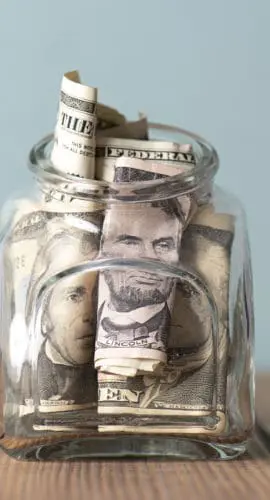 A glass jar filled with rolled and folded U.S. dollar bills sits on a wooden surface against a plain, light-colored background.