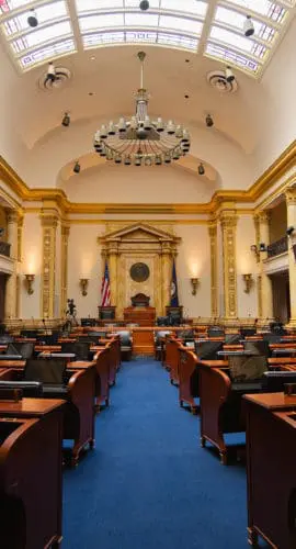 A legislative assembly hall with empty desks and a large chandelier overhead.