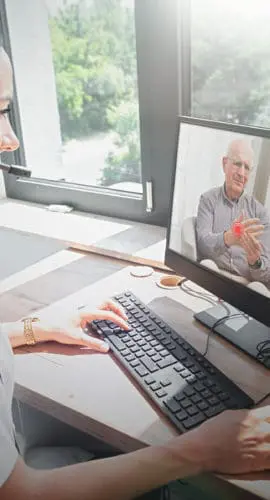 A woman wearing a headset is using a computer to have a video call with an older man who appears on the screen.