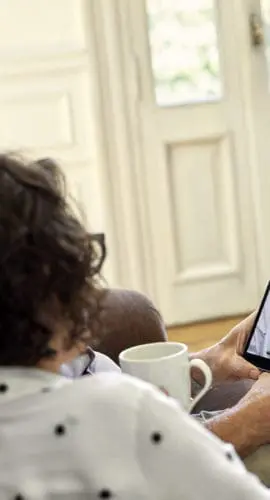 A couple sits on a couch having a video call on a tablet with a smiling woman.