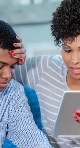 A woman is looking at a tablet while placing her hand on the forehead of a person sitting next to her.