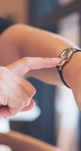 A person is pointing at their wristwatch while wearing a dark sleeve.