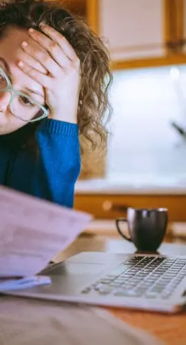 A person with curly hair and glasses appears stressed while looking at documents next to a laptop, a cup, and a glass of orange juice in a kitchen setting.