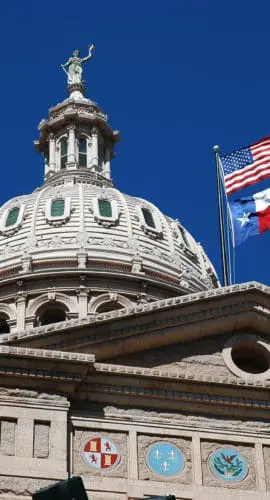 A grand architectural dome features a prominent American flag and another flag set against a clear blue sky.