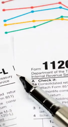 A calculator and a pen are placed on top of scattered IRS tax forms, including Form 1120 and 1120-L, with graphs in the background.