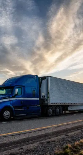 A blue semi-truck drives on a highway at sunset with a dramatic sky in the background.