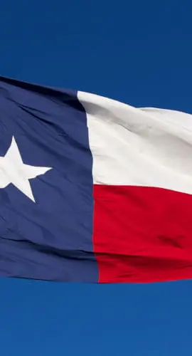 The Texas state flag, featuring a single white star on a blue vertical stripe next to horizontal white and red stripes, is shown flying against a clear blue sky.