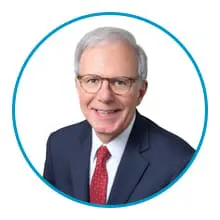 A person with white hair and glasses is smiling while wearing a dark suit and a red tie.
