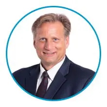 A person with light-colored hair is wearing a dark suit and tie, smiling, and is framed within a blue circle on a white background.