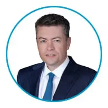 A person wearing a dark suit, white shirt, and blue tie is framed within a blue circular border.