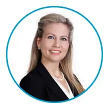 A woman with blonde hair, wearing a black suit and a white blouse, smiles against a white background, encircled by a blue border.