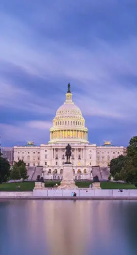 The U.S. Capitol building is lit up against a twilight sky with a reflection on a calm body of water in the foreground.