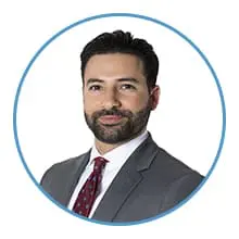 A man with a beard and short dark hair is wearing a grey suit, white shirt, and red tie, framed within a blue circle.