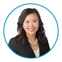 A smiling person with shoulder-length dark hair is wearing a black blazer, white top, and a pearl necklace, framed within a blue circle.