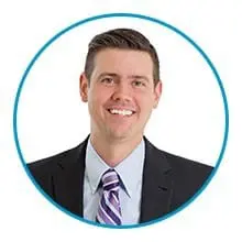 A person with short dark hair, wearing a suit, light blue dress shirt, and a purple striped tie, smiles against a white background within a blue circular frame.