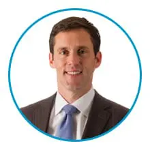 A man in a business suit with a light blue tie is smiling, set against a white background within a blue circular border.