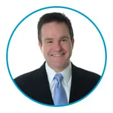 A person is smiling and wearing a suit with a light blue tie, framed by a blue circular border.