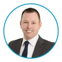 A man in a suit and tie smiles against a white background, framed by a blue circle.
