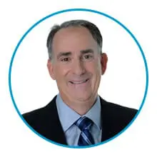 A smiling individual wearing a suit and tie is set against a white background within a circular blue border.