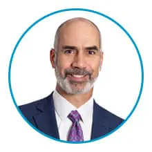 A man with a beard, wearing a suit and tie, is smiling with a white background framed by a blue circle.