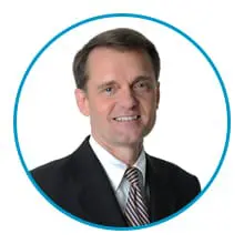 A person is wearing a dark suit, white shirt, and striped tie with a blue circular border around them.