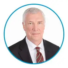 A person with short gray hair wearing a dark suit, white shirt, and striped tie is framed within a blue circle.