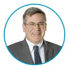 A man wearing glasses, a gray suit, and a blue tie is smiling against a white background within a blue circular frame.